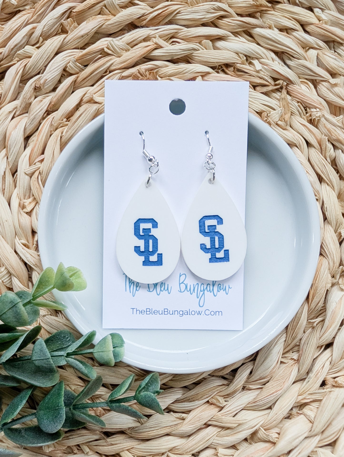 Pair of earrings with 'S' design on a white card with brand name, placed on a woven surface with green leaves.